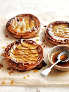 three pastries with apple slices on top and syrup in a bowl next to them