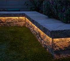 a stone wall with some lights on it in the grass near flowers and bushes at night