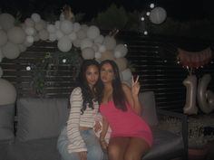 two women sitting on a couch with balloons in the air behind them and one pointing up