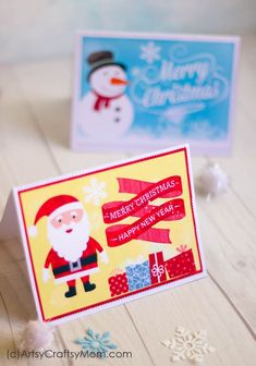 two christmas cards sitting on top of a wooden table next to snowflakes