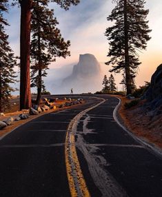 an empty road in the middle of some pine trees and mountains behind it with a person standing on one side