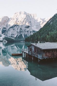 a small cabin sits on the edge of a lake with mountains in the background,