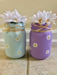 two painted mason jars with daisies tied to them on a tile floor next to a wall