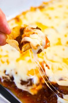 a hand holding a tortilla chip with cheese and sauce on it in front of a casserole dish