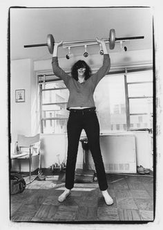 a woman standing on one leg holding two dumbbells above her head and looking at the camera