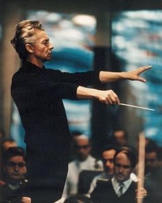 a man in black shirt and white pants holding a conductor's baton while people watch