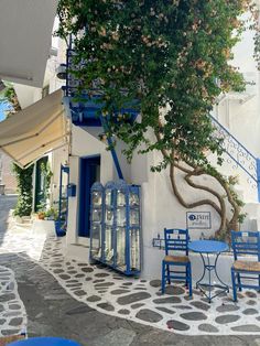 the outside of a restaurant with blue chairs and tables under a tree in front of it