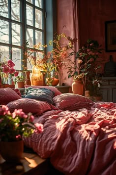 a bed with pink sheets and pillows in front of a window filled with potted plants