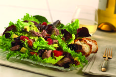 a salad with meat and fruit is on a plate next to a fork, knife and glass of wine