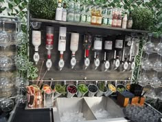 an outdoor bar with lots of bottles and glasses on the wall above it, surrounded by plants