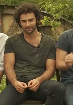 three men are sitting on a bench in front of some trees and bushes, one is looking at the camera