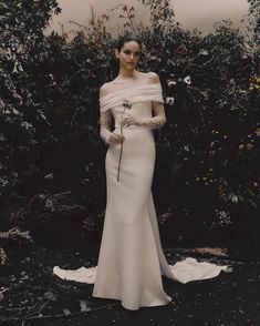 a woman standing in front of some bushes wearing a white dress and holding a flower