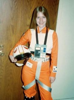 a woman dressed in an orange astronaut suit holding a baseball glove and helmet while standing next to a door