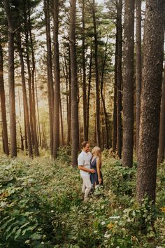 a man and woman standing in the middle of a forest with tall trees behind them