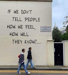 two people walking past a building with graffiti on the wall that says if we don't tell people how we feel, how will they know?