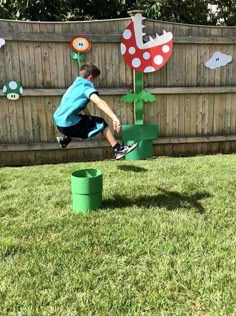 a young boy jumping over two green buckets on top of grass in front of a wooden fence