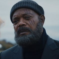 an older man wearing a beanie stares into the distance while standing next to a woman