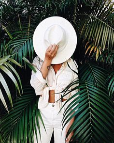 a woman wearing a white hat standing in front of palm trees and plants with her hands on her face