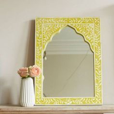 a yellow mirror sitting on top of a wooden shelf next to a vase with flowers