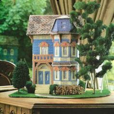 a doll house on a table with trees and bushes in the foreground, next to a window