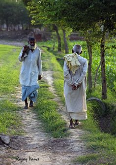 two men walking down a dirt road next to trees