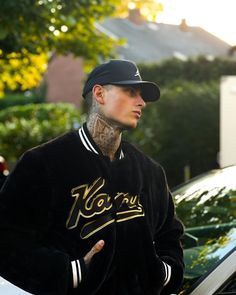 a man with tattoos standing in front of a white car wearing a black baseball cap