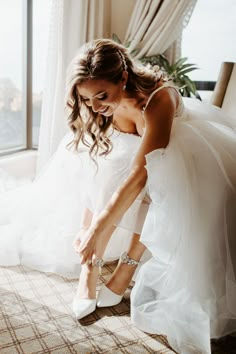 a woman in a white dress is tying her wedding shoes on the floor near a window