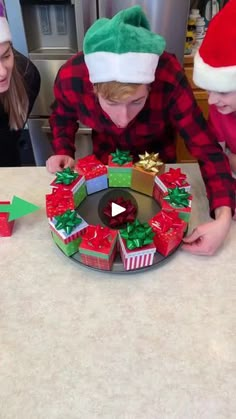 three people in christmas hats are looking at a paper wreath on a table with presents