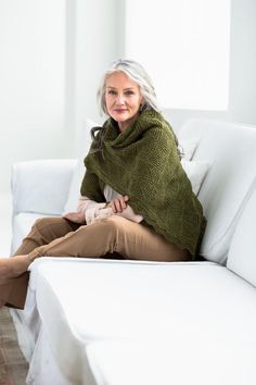 an older woman sitting on a white couch wearing a green shawl and tan pants