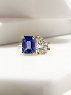 an engagement ring with two pear shaped diamonds and a blue sapphire in the center, sitting on a white surface