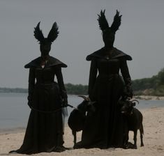 two people in black costumes standing on the beach with their goats wearing horns and headgear