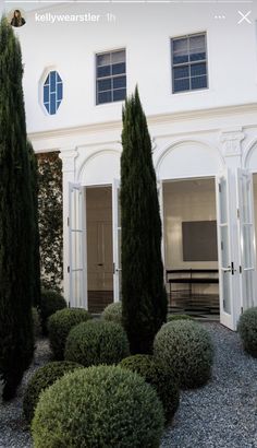 a house with several trees and bushes in front of the door, on gravel ground