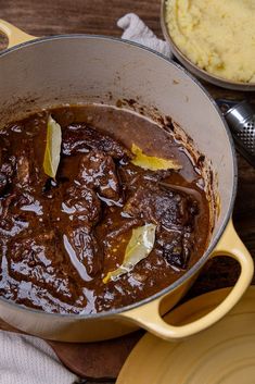 a pot full of stew sitting on top of a wooden table next to a bowl with mashed potatoes