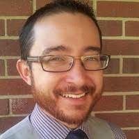 a man wearing glasses and a tie standing in front of a brick wall smiling at the camera