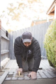 a man kneeling down on top of a tile floor next to a wooden fence and shrubbery