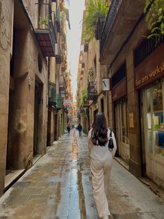 a woman walking down a wet street in an alley way with buildings on both sides