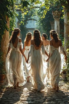 four women in white dresses walking down a stone path with greenery on either side