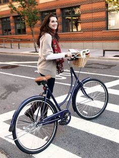 a woman is riding her bike down the street while wearing a scarf and holding a basket