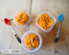 three plastic containers filled with food sitting on top of a wooden table next to two spoons