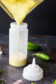 a person pouring mustard into a jar with peppers around it and on the table next to them