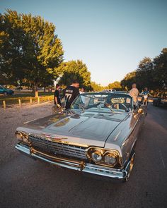 an old car is parked on the side of the road with people standing around it