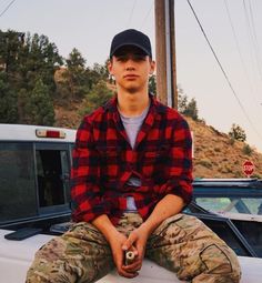 a young man sitting on the hood of a car wearing camo pants and a baseball cap