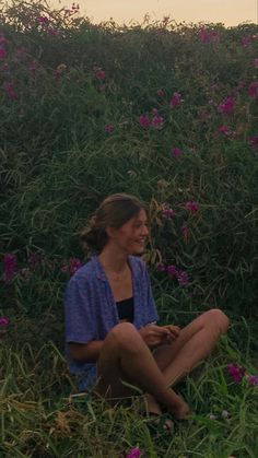 a woman sitting on the ground in front of some purple flowers and smiling for the camera