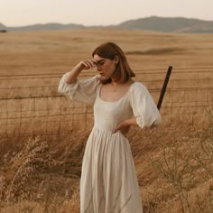 a woman in a white dress is standing by a fence and looking off into the distance