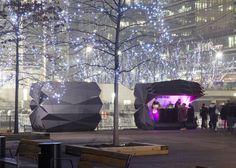 people are standing around in an open area with lights on the trees and buildings behind them