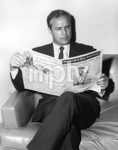 a man in a suit and tie sitting on a couch reading a newspaper with his legs crossed