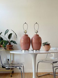 two pink vases sitting on top of a white table next to a chair and potted plant