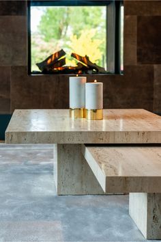 two candles sitting on top of a wooden table in front of a window with a fireplace