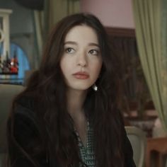 a woman with long dark hair sitting in a chair and looking at the camera while wearing earrings