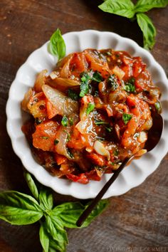 a white bowl filled with pasta and vegetables on top of a wooden table next to basil leaves